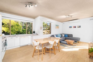 A modern kitchen with white cabinets, a dining area with a wooden table and chairs, a grey sofa, and a large window overlooking greenery.