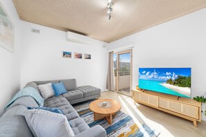A living room with a grey sofa, a coffee table, and a TV displaying a beach scene. - Del Rae 15 Shoal Bay Apartment (Shoal Bay)