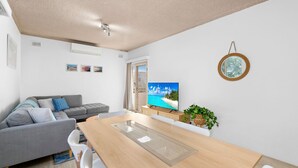Una sala de estar moderna con un sofá gris, una mesa de comedor de madera y un televisor que muestra una escena de playa tropical.