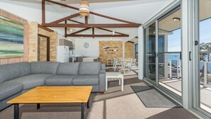 Ein modernes Wohnzimmer mit einer grauen Eckcouch, einem Holztisch und einem Essbereich mit weißen Stühlen. Im Hintergrund ist eine Küche erkennbar, und ein großes Glasfenster führt auf einen Balkon mit Blick aufs Wasser.