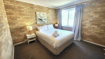 A hotel room with a brick wall, a bed with white linens, a nightstand with a lamp, and a window with curtains.
