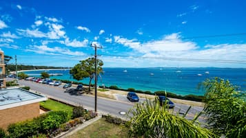 A coastal road with parked cars, a clear blue sky, and boats on the water.