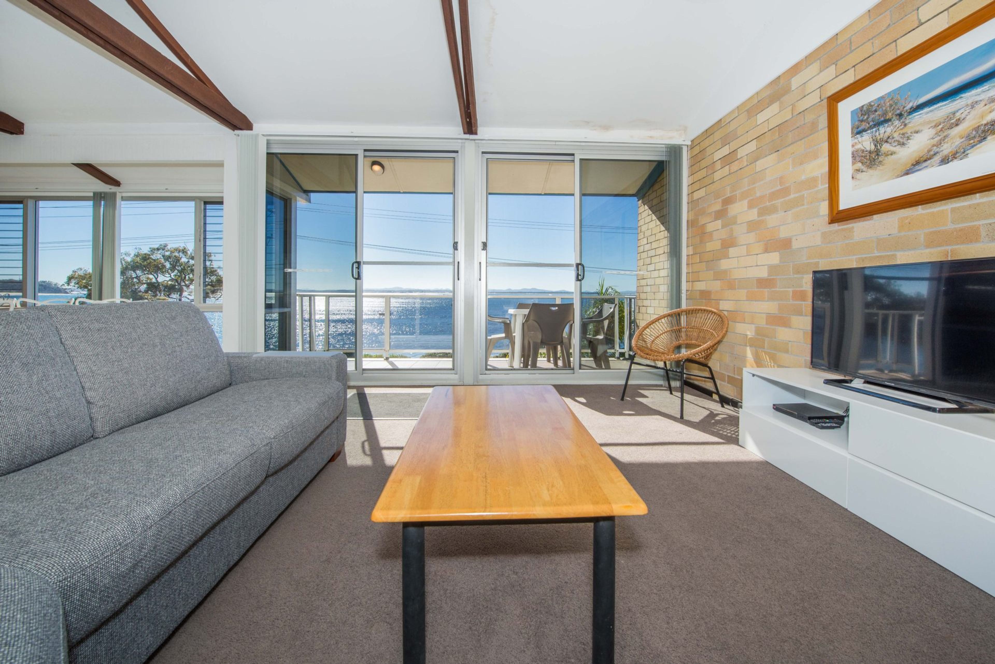 A modern living room with a sofa, a wooden table, a TV, and a balcony with a view.
