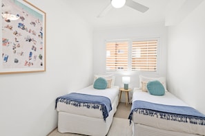 Een slaapkamer met twee bedden, een plafondventilator, een raam met zonwering en een aan de muur hangend schilderij.