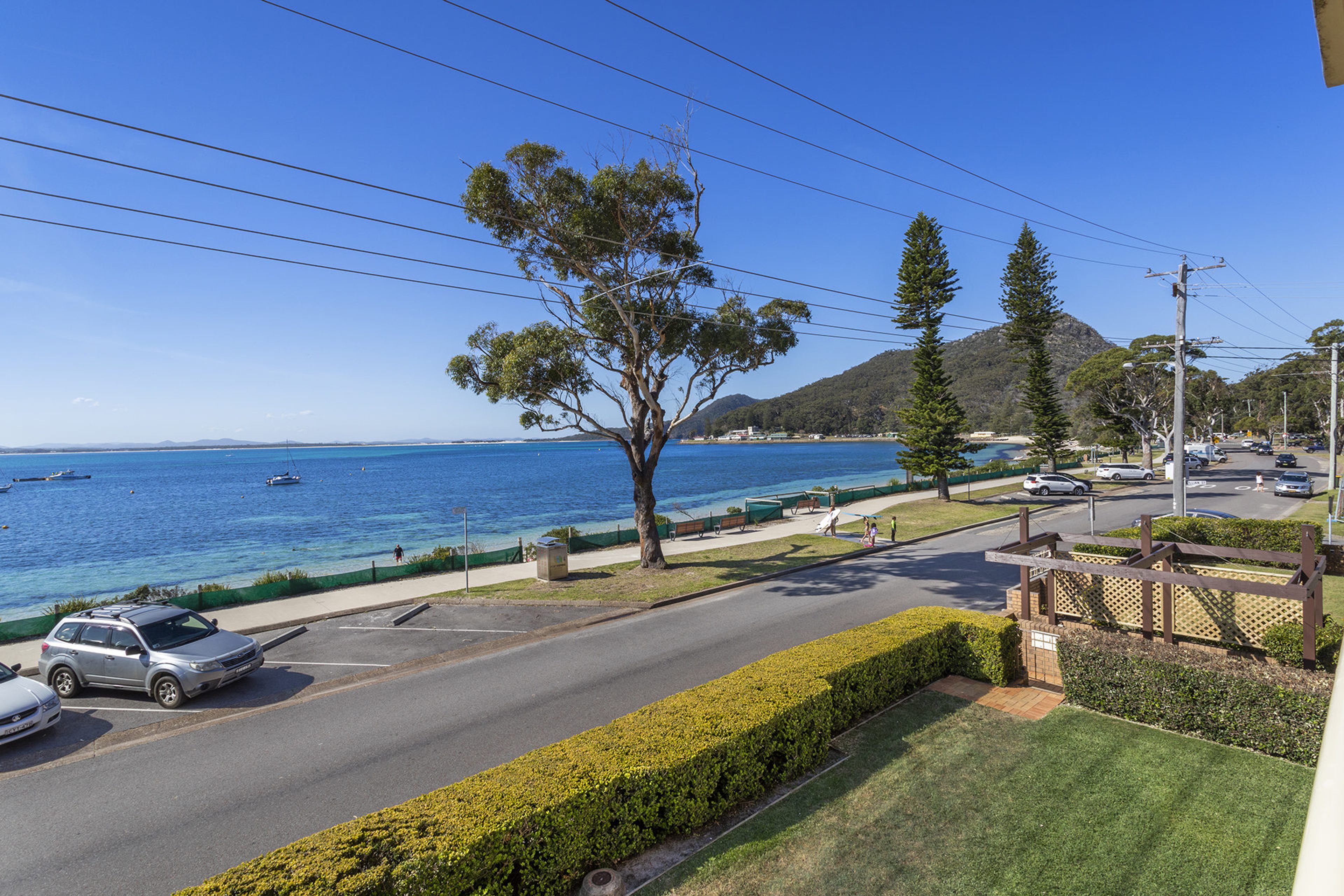 A coastal road with a view of the sea, a tree-lined sidewalk, and parked cars.