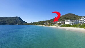 A coastal hotel with a beach view, a mountain in the background, and a red logo. - Three bedroom unit in Shoal Bay (Shoal Bay)