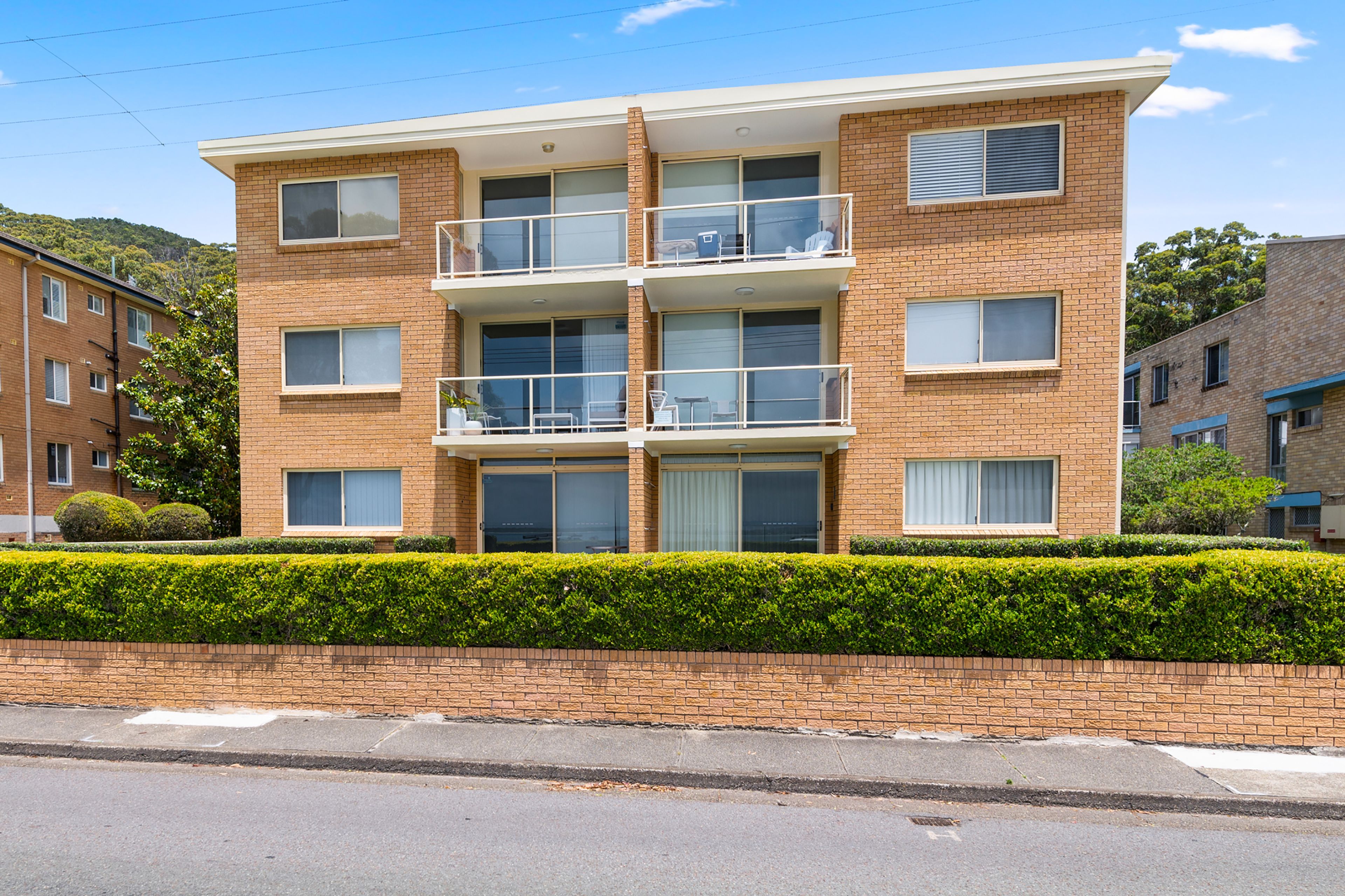 A multi-story residential building with a brick facade, balconies, and a well-maintained hedge in front.