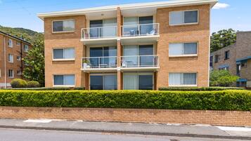 A multi-story residential building with a brick facade, balconies, and a well-maintained hedge in front.