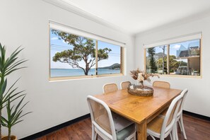 Un coin repas avec une table en bois, des chaises blanches et une vue sur la mer grâce à de grandes fenêtres.