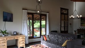 A cozy living room with a wooden desk, a sofa, and a coffee table. There is a TV mounted on the wall, a window with curtains, and a chandelier.