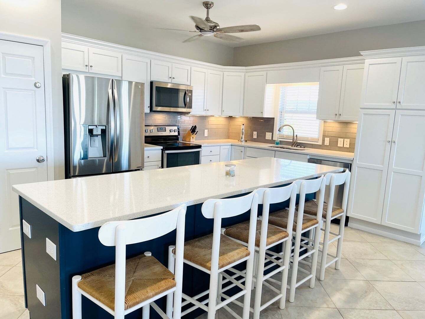 A modern kitchen with white cabinets, stainless steel appliances, a central island with bar stools, and a ceiling fan.