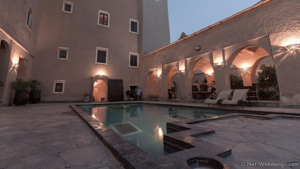 Un área de piscina con suelo de piedra, una piscina y sillas de descanso. Se observan paseos con arcos y un edificio con varias ventanas.