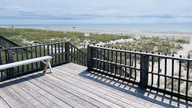 A wooden deck with a bench, overlooking a beach with sand dunes and a fence. - Seaside: Spacious 4BR oceanfront home in Holden Beach, perfect for 12 guests. (Holden Beach)
