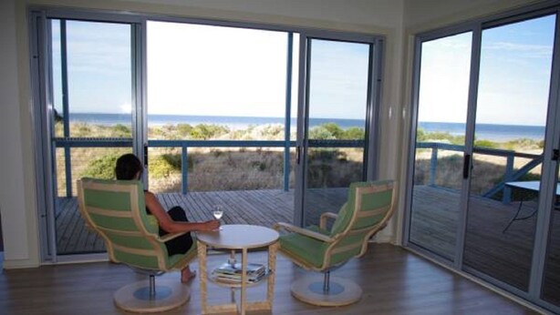 Ett rum med stora fönster som ger utsikt över stranden. Det finns två gröna stolar och ett litet bord.