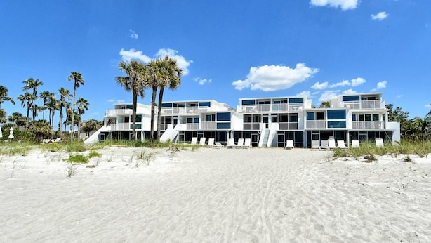 Un hôtel situé en bord de mer, doté de nombreux balcons, avec une plage de sable en premier plan et des palmiers.