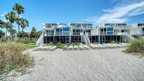 Un hôtel moderne situé en bord de mer, avec plusieurs étages, de grandes fenêtres et une plage de sable où l’on trouve des chaises longues.