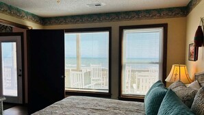 Un dormitorio con una cama grande, dos ventanas con persianas, un ventilador de techo y vistas a un paisaje urbano.