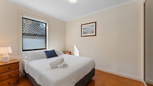 Un dormitorio con cama, ventana con mosquitera, mesita de noche con lámpara, un cuadro enmarcado en la pared y un aparador de madera.