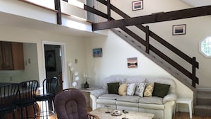 A spacious living area with a high ceiling, a wooden staircase, a sofa set, a dining table with chairs, and a decorative bird sculpture.