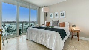 Une chambre moderne avec un grand lit, une table de chevet, une lampe et une vue dégagée sur l’extérieur grâce à une grande fenêtre.