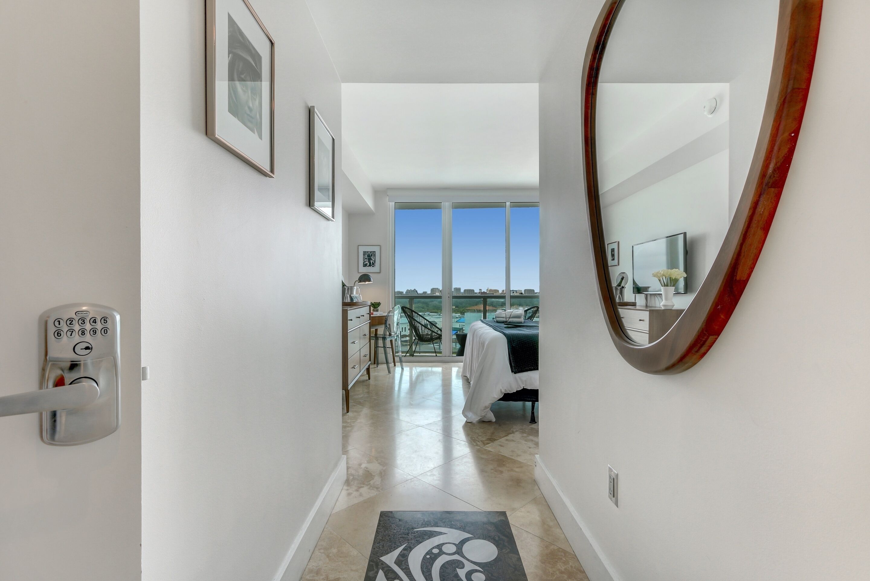 A hallway leading to a bedroom with a large window, a bed, and a desk.