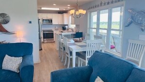 A modern kitchen with white cabinets, a dining area with white chairs, and a large window with a view. - Libby's Lookout: Spacious 4-bedroom retreat near the beach in Holden Beach, NC. (Holden Beach)