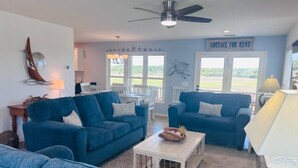A living room with two blue sofas, a coffee table, and a ceiling fan. There is a sign that reads "COTTAGE FOR RENT" on the wall. - Libby's Lookout: Spacious 4-bedroom retreat near the beach in Holden Beach, NC. (Holden Beach)