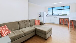 A modern living room with a beige sectional sofa, a small white side table, and a red patterned pillow. A kitchen area is visible with white cabinets, a refrigerator, and a window with a view of buildings. - Blue Door Retreat  - Excellent Off Peak Special!  Nightly rates reduced!! (Edithburgh)