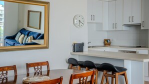 A modern kitchen with white cabinets, a built-in oven, and a dining area with wooden chairs and a table. - Blue Waters Unit 8/59 Ronald Ave, Shoal Bay - This cosy unit has the views and the position! (Shoal Bay)