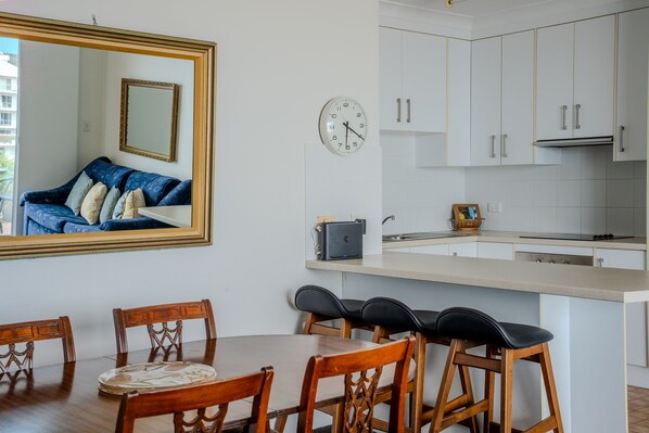 A modern kitchen with white cabinets, a round clock, a dining area with wooden chairs, and a large mirror on the wall. - Blue Waters 8 Shoal Bay Apartment (Shoal Bay)