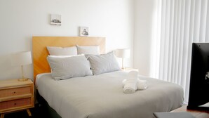 A hotel room with a bed, two bedside tables, a TV, and a window with blinds. - Blue Waters Unit 8/59 Ronald Ave, Shoal Bay - This cosy unit has the views and the position! (Shoal Bay)