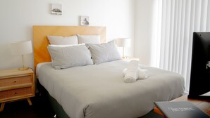 Un lit bien fait, avec une tête de lit, deux tables de chevet avec des lampes, une télévision et une petite œuvre d’art accrochée au mur.