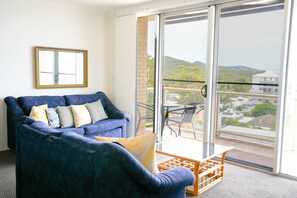 A room with a blue sofa, a small table, and a chair. There is a large window with a view of buildings and hills. - Blue Waters 8 Shoal Bay Apartment (Shoal Bay)