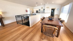 Un comedor amplio con mesa de madera y sillas de mimbre, una cocina al fondo y una escalera con barandilla negra.