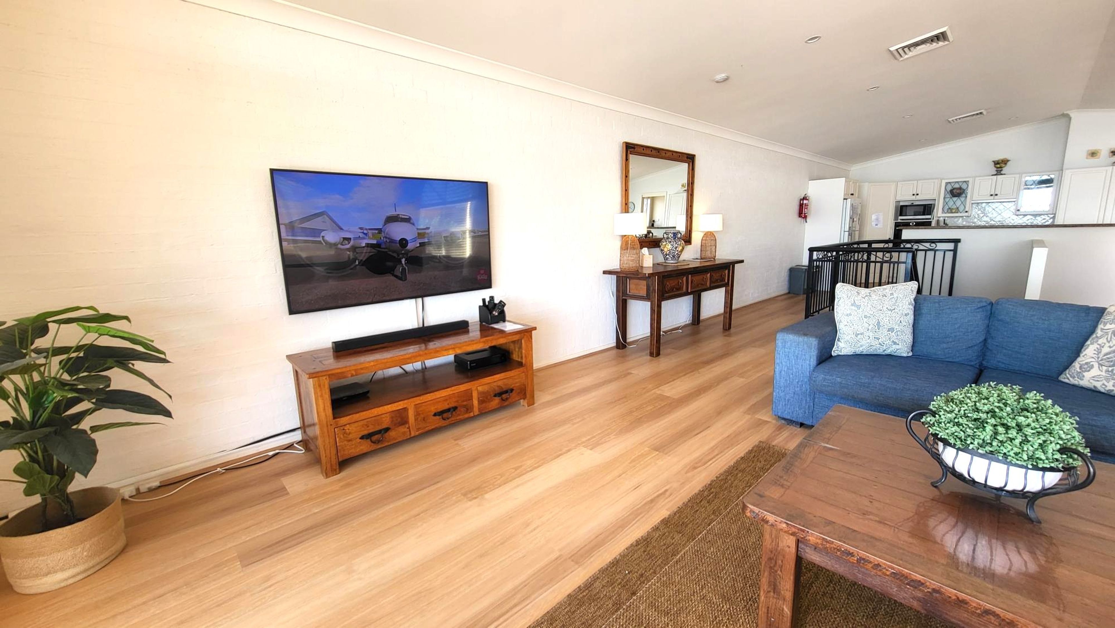 A living room with a flat-screen TV, wooden entertainment unit, blue sofa, and a kitchen area in the background.