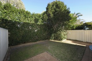 A backyard with a grassy lawn, a brick patio, and a tall tree. - Marine Stunning Waterfront Home in Fingal Bay (Fingal Bay)