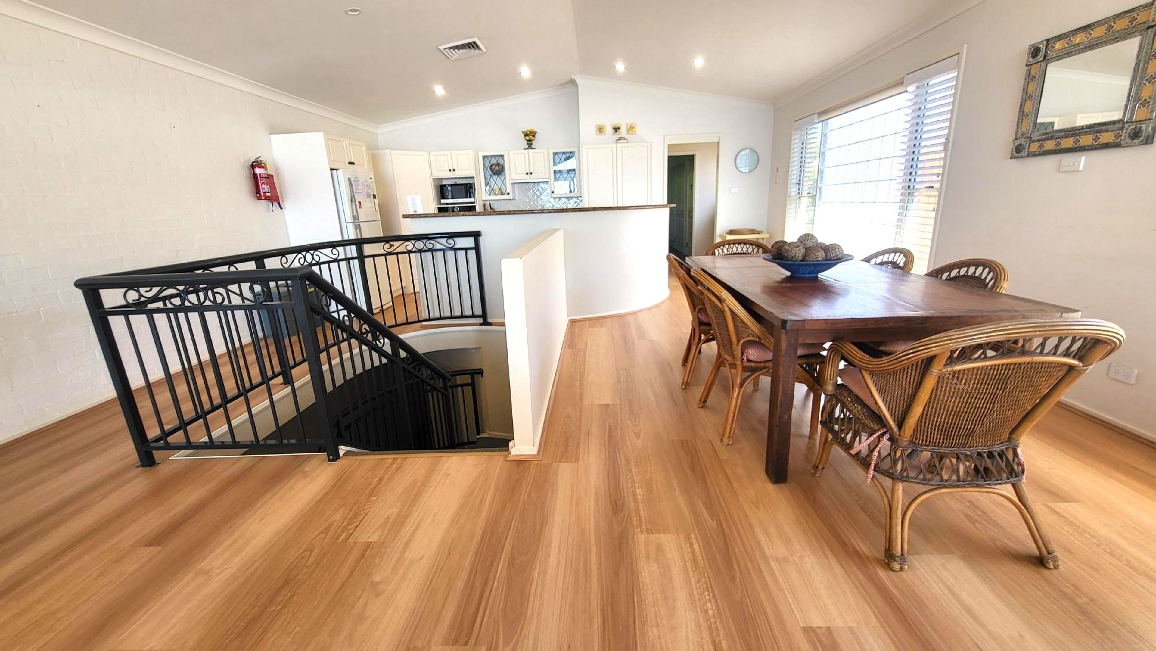 A dining area with a wooden table and wicker chairs, a kitchen with white cabinets, and a staircase with a black railing.