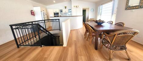 A dining area with a wooden table and wicker chairs, a kitchen with white cabinets, and a staircase with a black railing.