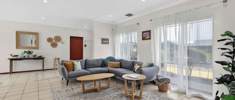 Una sala de estar moderna con un sofá gris, dos mesitas de madera, un tapete estampado y un ventanal con cortinas traslúcidas.