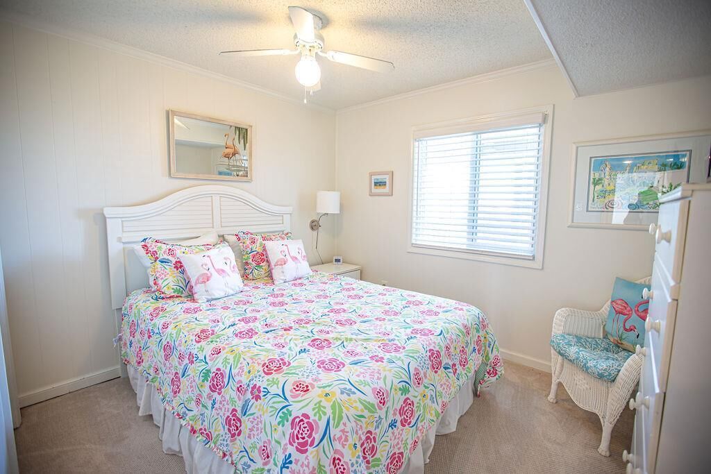 A bedroom with a large bed, a ceiling fan, a window with blinds, and a chair.