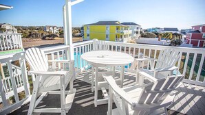 A covered outdoor area with white Adirondack chairs and a round table, overlooking a pool and colorful buildings. - Relax and unwind in Feelin' Beachy (Holden Beach)
