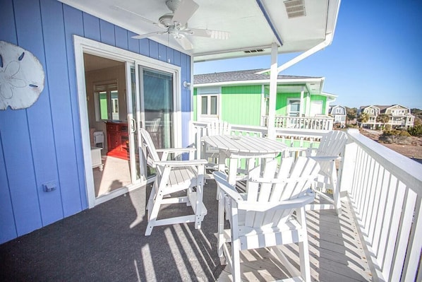 A balcony with white Adirondack chairs and a table, overlooking a green house and beach.