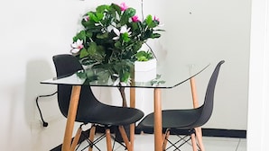 Una pequeña sala de reuniones con una mesa de vidrio, dos sillas negras y una planta en maceta con flores rosadas.