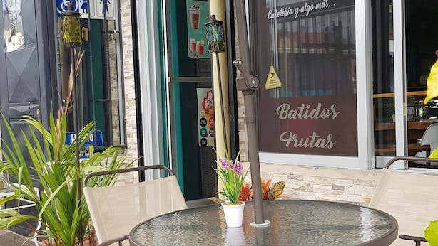 Un área de estar al aire libre con una mesa redonda y sillas, plantas en macetas y un escaparate con un letrero que dice "Batidos Frutas".