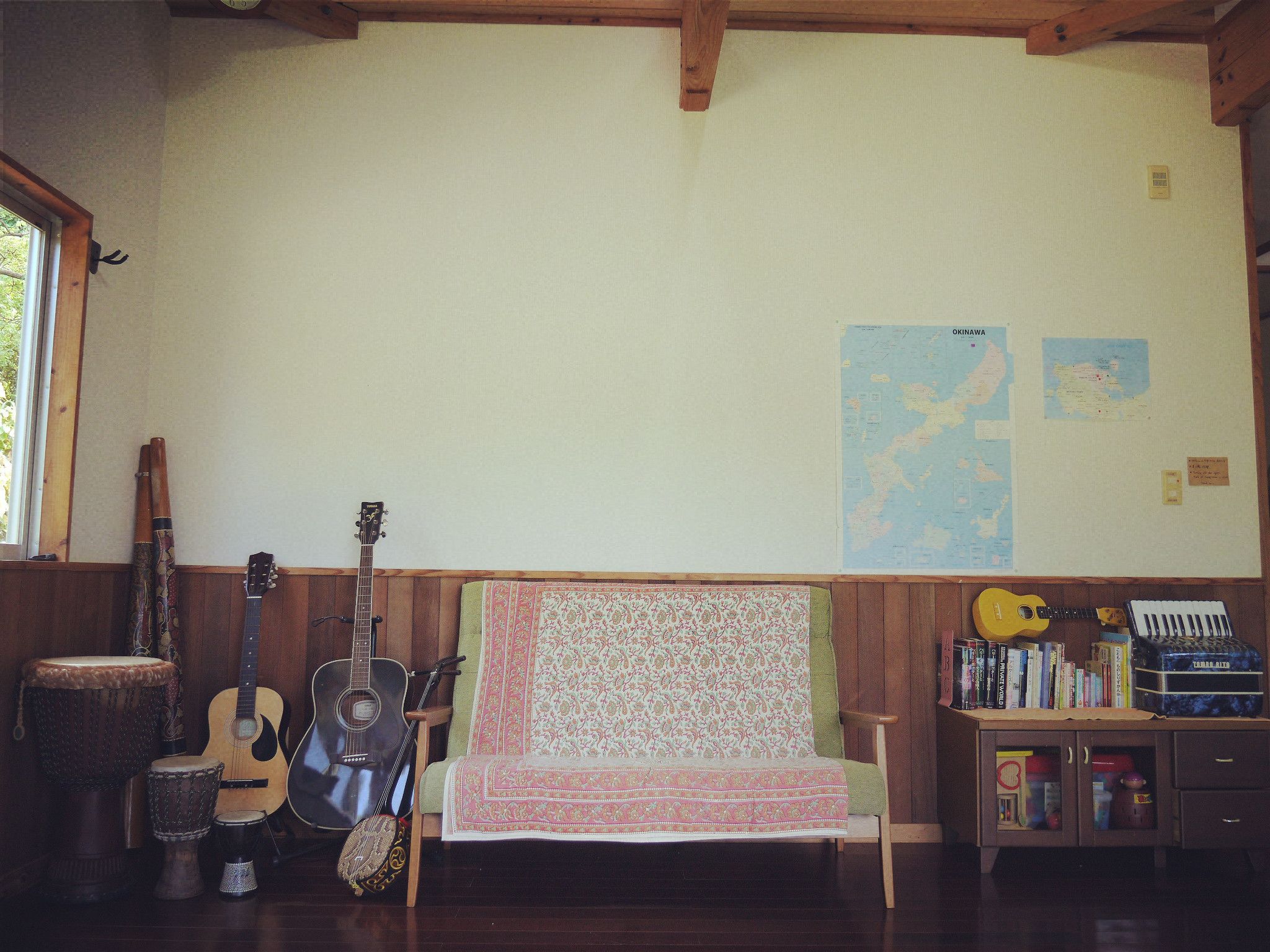 A room with a wooden bench, a guitar, a tambourine, a drum, and a keyboard. There are maps on the wall and a cabinet with books and a suitcase.