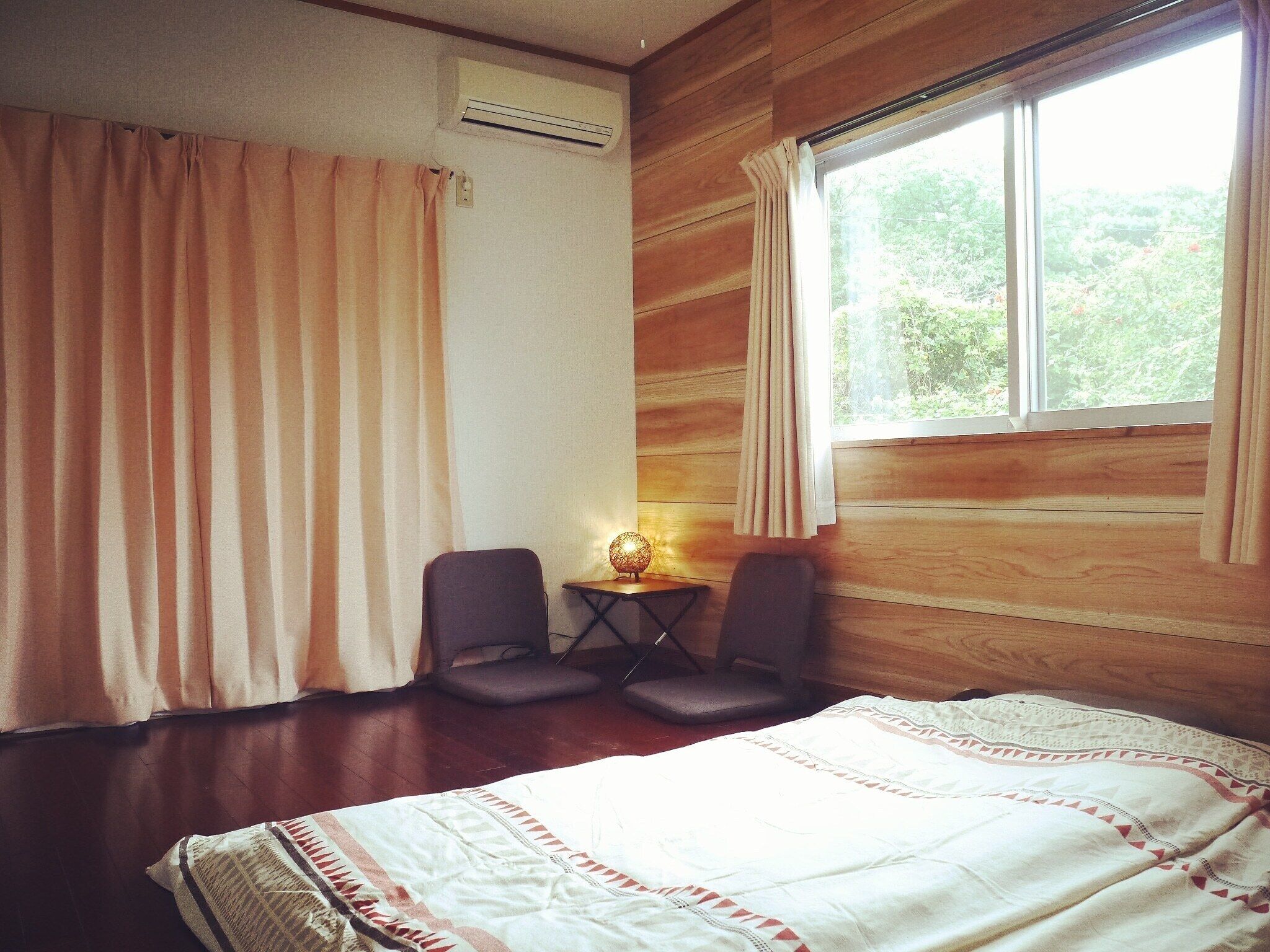 Een kamer met een bed, twee stoelen, een kleine tafel met een lamp, een airconditioning en een raam met gordijnen.