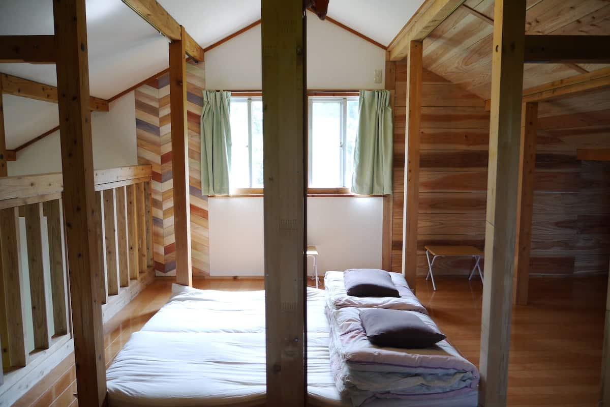 Une cabane avec une chambre à coucher aménagée en mezzanine, des murs en bois, une fenêtre avec des rideaux et un lit avec des oreillers.