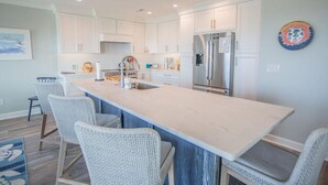 A modern kitchen with white cabinets, a large island, stainless steel appliances, and wicker chairs. - Oceanfront oasis in Holden Beach! Spacious 6BR, 6BA, sleeps 14. (Holden Beach)