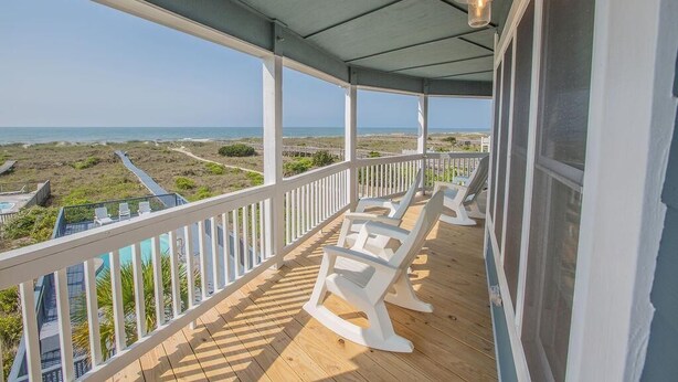 A covered porch with white rocking chairs, a wooden deck, and a view of the beach and ocean. - Oceanfront oasis in Holden Beach! Spacious 6BR, 6BA, sleeps 14. (Holden Beach)