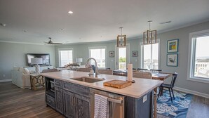 A modern kitchen with a central island, a dining area with a table and chairs, and a living room with a sofa and TV. - Oceanfront oasis in Holden Beach! Spacious 6BR, 6BA, sleeps 14. (Holden Beach)